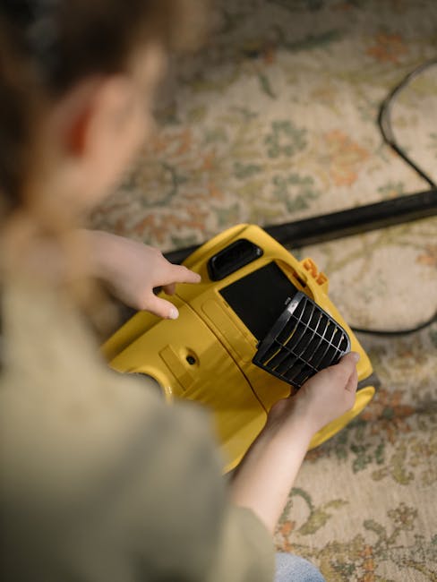 Close-up of a professional wet vacuum cleaner with a transparent dust collection chamber filled with water and foam, positioned on a pinkish-beige carpet in a room with white walls and wooden skirting